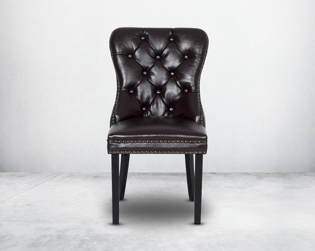 Dining chair upholstered in faux leather with front view buttoned tufted back the chair has antique gold rivets, legs in dark vintage brown wood.
